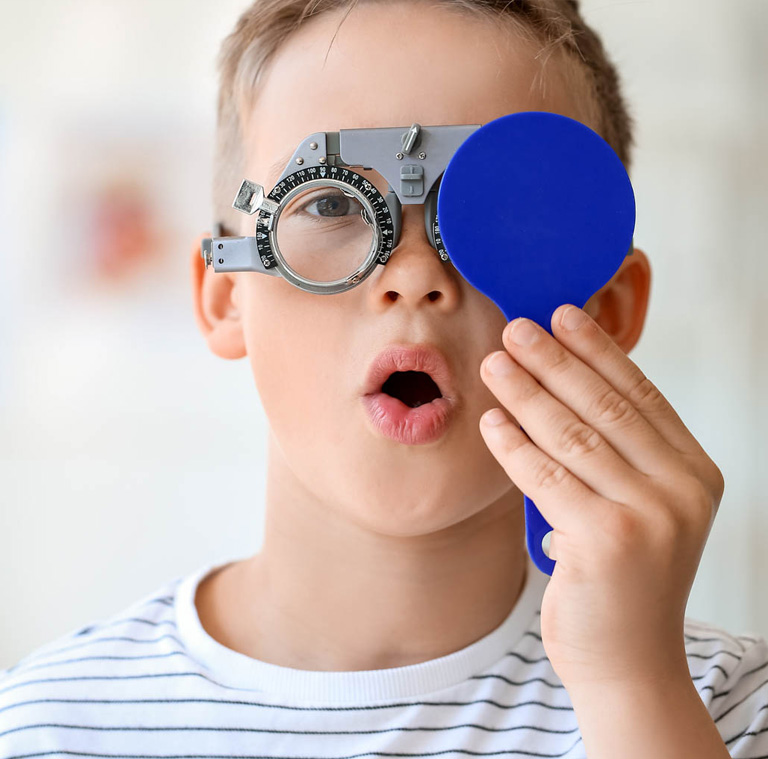 A boy wearing eye test glasses holding a blue paddle to his eye.